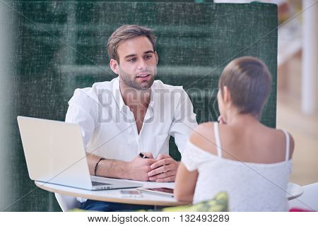 Knowledgable male tutor busy mentoring his new bright female student, image taken through glass with reflections visible as well as a host of technology on the table between them.