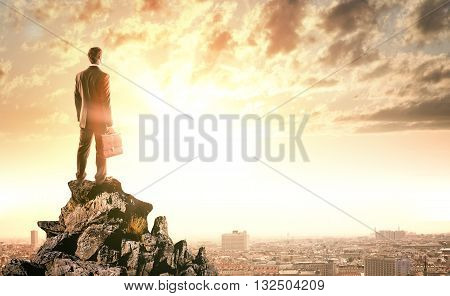 Young businessman standing on edge of rock mountain and looking at city