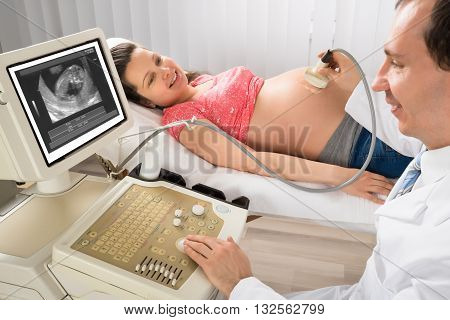 Happy Male Doctor Moving Ultrasound Transducer On Pregnant Woman's Belly While Looking At Screen
