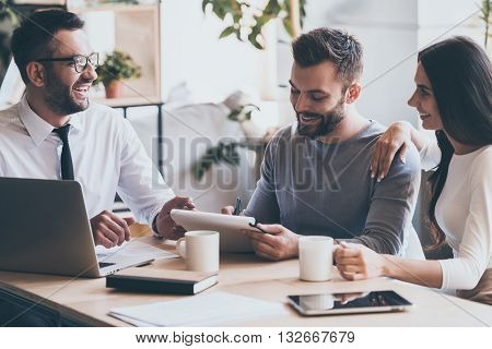 Just put your signature here! Cheerful young man signing some documents while sitting together with his wife and man in shirt and tie