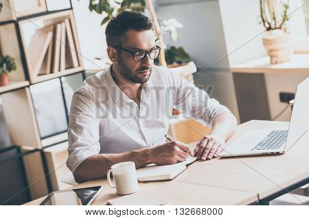 Concentrated at work. Confident mature man writing something in note pad while sitting at his working place in office