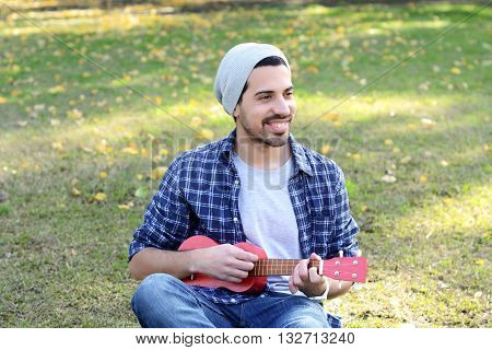 Portrait of young handsome man playing the ukelele in a park. Outdoors.