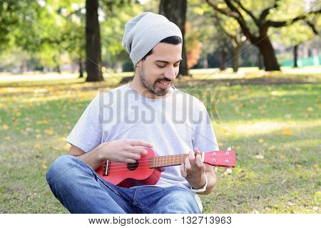 Portrait of young handsome man playing the ukelele in a park. Outdoors.