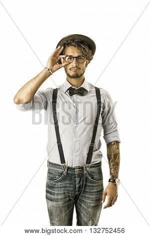 Portrait of brunette young man in glasses, hat, bow-tie, suspenders and shirt looking at camera. Studio shot, isolated on white