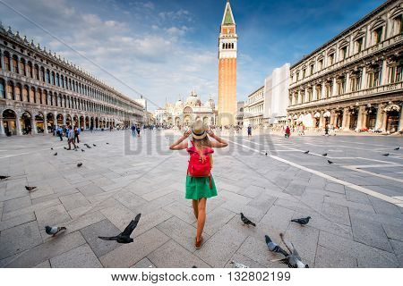 Young female traveler with hat and backpack standing on San Marco square with tower and basilica on the background in Venice. Back view with copy space