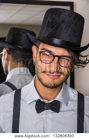Portrait of brunette young man in glasses, hat, bow-tie, suspenders and shirt looking at camera. Mirror behind him