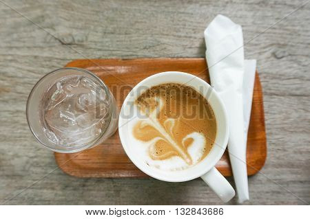 Set of hot coffee with streamed milk and a light layer of foam on wooden tray served with ice water and a piece of napkin