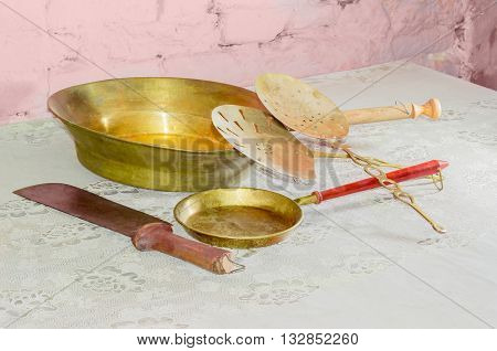 Some subjects of a older kitchen utensils consisting of two various copper frying pans and slotted spoons large kitchen knife on table with old tablecloth against background of the brickwork