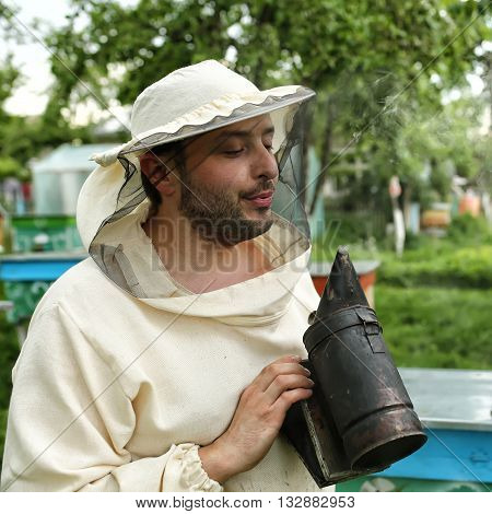 beekeeper in protective clothing holding smoker while standing at apiary