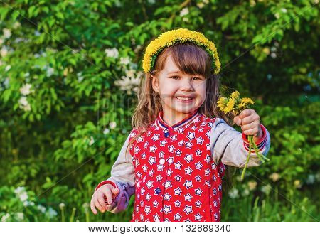 Joyful girl with a wreath of dandelions on her head and a bouquet of dandelions in hand on background of green tree in summer Park