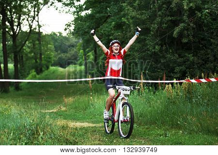 Crossing the line. Winner. Victory moment. Young red-haired woman cyclist in protective helmet crossing the finish line breaking the tape. Win the cycling race. Bicycle sport.