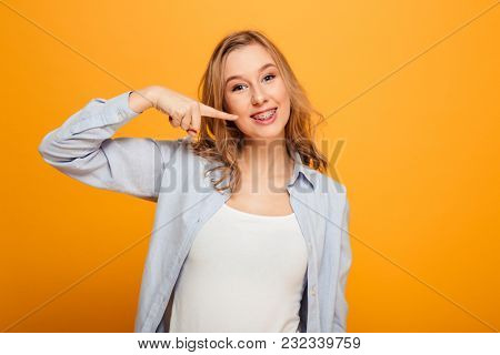 Portrait of a smiling young girl with braces pointing finger at her mouth isolated over yellow background