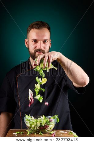 Portrait Of A Handsome Mischevious Male Chef Cook In Black Uniform Making Delicious Vegetable Salad 