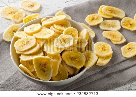 Heap Of Dried Banana Chips Snack In White Bowl On Rustic Table With Grey Napkin
