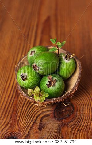 Green Feijoa In A Coconut Shell Hulf On A Wooden Background. Ripe Tropical Fruits And Mint Leaves, P