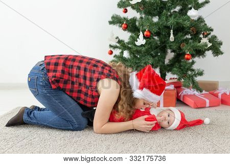 Holidays And Christmas Concept - Young Mother With Baby Santa Claus In Front Of Xmas Tree