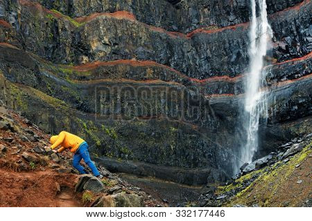 Tourist in front of Hengifoss canyon and waterfall, the third highest waterfall in Iceland, surrounded by basaltic strata with red layers of clay between the basaltic layers.