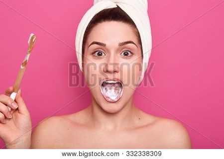 Shot Of Adorable Female With White Towel On Head And Nacked Shoulders, Brushing Her Teeth, Looking W