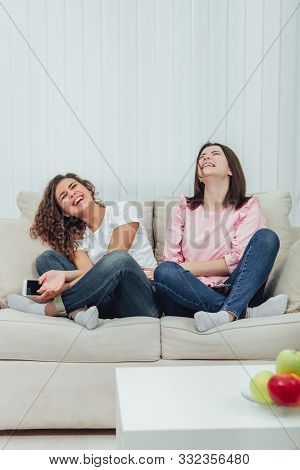 Two Attractive Girlfriends Sitting On The Sofa In Lotos Position, With Smartphones In Hands, Smiling