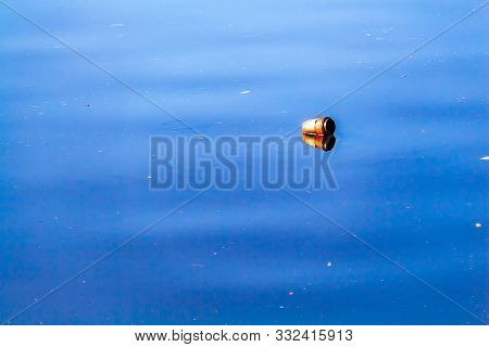 A Coffee Cup With A Plastic Lid Lies On Its Side, Floating In The Water Of A River. The Water Reflec
