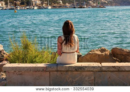 Young Attractive Woman Sitting On A Wall At The Mediterranean Sea Looking At The Harbor.