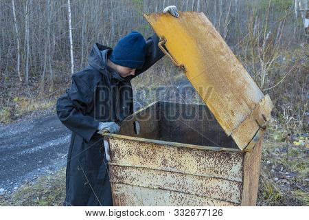 Homeless Woman In Black Coat And Blue Hat Is Open Street Garbage Cans. Woman Is Rummaging For Some F