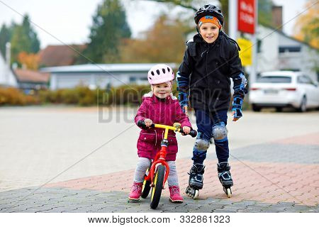 Kid Boy And Little Toddler Girl Playing Together Outdoors. Brother In Safety Clothes Skating With Ro