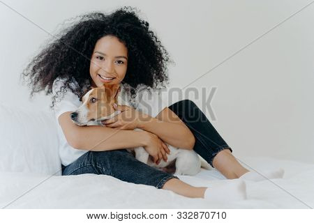 Photo Of Lovely Young Female Pet Owner Poses In Clean White Bedroom, Hugs Dog, Plays With Best Frien
