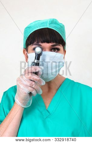 Woman Medic Holding Otoscope To Eye. Wearing Face Mask, Hair Net And Green Scrubs. Hand In Clear Lat