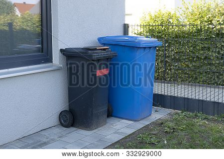 Trash Cans Of Black And Blue On Wheels Stand Near The Wall. Dirty Plastic Mobile Garbage Bins And Ho