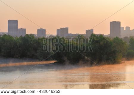 Houses And Trees On The River Bank. Tall Houses, Lush Trees On The Riverbank At Dawn. Fog Over The W