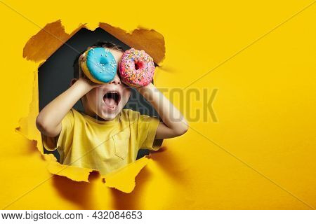Happy cute boy is having fun played with donuts on Yellow background wall. Bright photo of a boy. Colored donuts