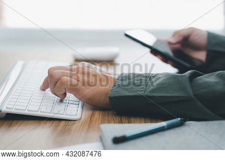 Man's Hand Typing On Desktop Keyboard On Wood Desk With Holding Smartphone Business Working In Offic