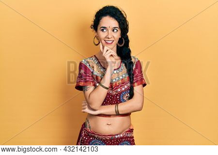 Young woman wearing bindi and bollywood clothing with hand on chin thinking about question, pensive expression. smiling with thoughtful face. doubt concept. 