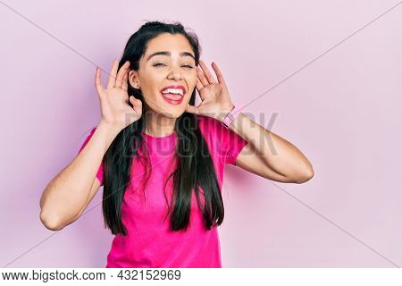 Young hispanic girl wearing casual pink t shirt trying to hear both hands on ear gesture, curious for gossip. hearing problem, deaf 