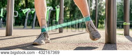 Closeup front view of legs feets of man guy training with elastic rubber band at the stadium outdoors. Athlete adult male person during active workout with additional sport equipmant in summertime