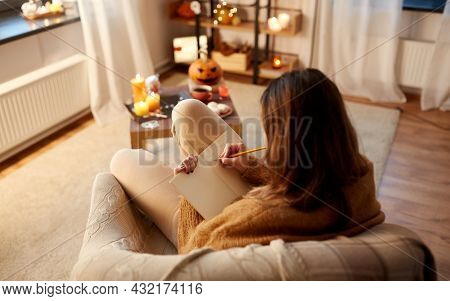 halloween, holidays and leisure concept - young woman with pencil writing to diary at cozy home