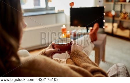 halloween, holidays and leisure concept - young woman watching tv and drinking hot chocolate with her feet on table at cozy home