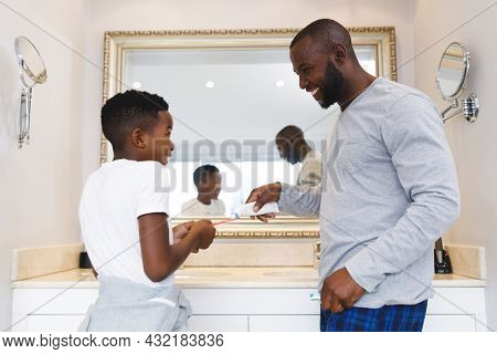 Smiling african american father and son putting toothpaste on toothbrush in bathroom. family spending time at home.