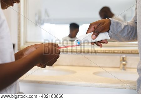 African american father and son putting toothpaste on toothbrush in bathroom. family spending time at home.