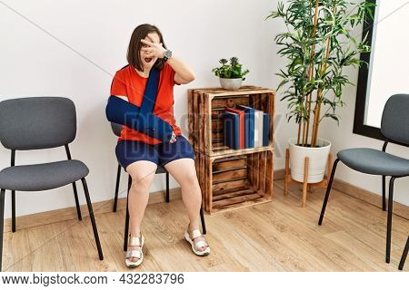 Young down syndrome woman sitting at doctor waiting room with arm injury peeking in shock covering face and eyes with hand, looking through fingers with embarrassed expression. 