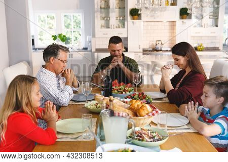 Caucasian grandfather and parents with son and daughter sitting at table and praying before dinner. family enjoying traditional mealtime together at home.