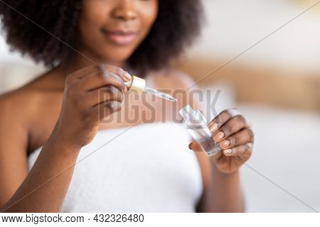 Unrecognizable Young Black Woman In Bath Towel Hlding Bottle Of Face Serum With Pipette Indoors, Clo