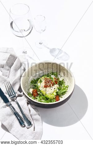 Avocado and stracciatella cheese salad. Fancy dinning with Italian salad bowl on white table with simple contemporary decor. Sunlight and harsh shadow still life. Avocado, cheese and tomato salad