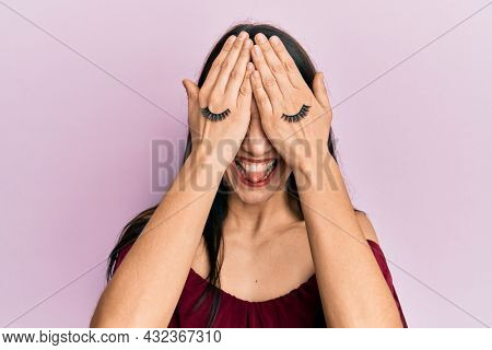 Young hispanic woman covering eyes with hands and fake lashes sticking tongue out happy with funny expression. 
