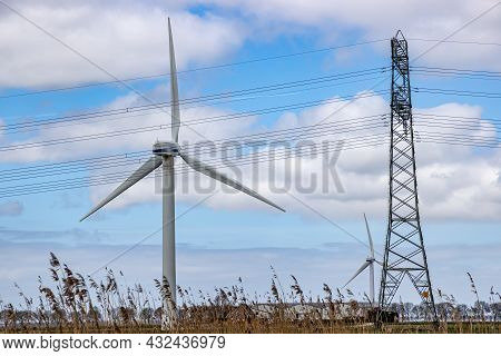 Huge White Metal Windmill Next To Power Tower With High Voltage Cables On Farm Agricultural Land, Bl
