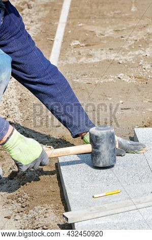 A Worker On A Sunny Day Flattens Granite Tiles With A Rubber Mallet While Assembling A New Sidewalk 