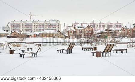 Winter City Landscape. Snow-covered Benches On The River Bank Against The Background Of Low-rise Bui