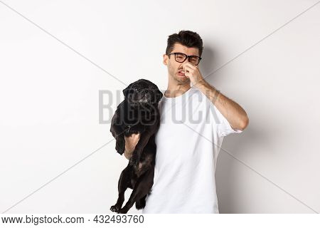 Young Man Shut Nose As Holding Pug, Disgusted With Bad Smell Fart Of Animal, Standing Over White Bac