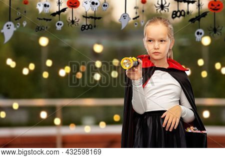 halloween, holiday and trick-or-treating concept - girl in black dracula cape or costume with candies and flashlight over roof top party background